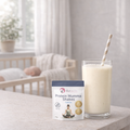 Glass of protein shake with a container labeled 'Protein Mumma Shakes' on a surface, with a blurred background of a room.